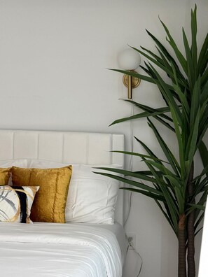 A bed with white bedding and gold and white pillows, a wall-mounted light fixture, and a large green plant. - 1BR Apt • 2 Queen Beds • Free Parking • Stair-Free Access • Near NYC (White Plains)