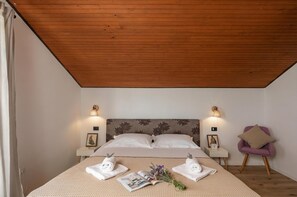 Une chambre avec un plafond en bois, un lit recouvert de draps blancs, deux tables de chevet avec des lampes, un fauteuil violet et un bouquet de fleurs sur le lit.