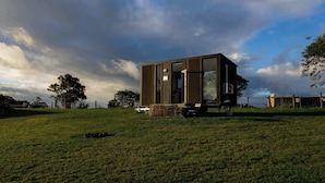 A modern, standalone building with a large glass window, situated on a grassy field with a view of the sky and trees. - Hills and Herds by Tiny Away (Shannon Brook)