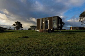 A modern, standalone building with a large glass window, situated on a grassy field with a view of the sky and trees.