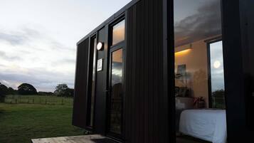 A small, dark wooden cabin with a bed, a table, and a view of the sky.