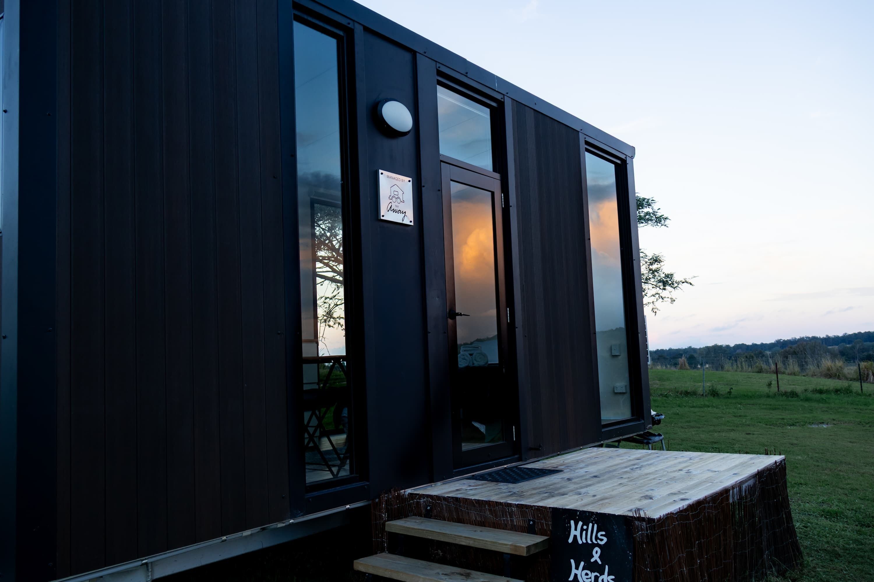 A modern, dark-colored building with a wooden bench outside, a sign reading 'Hills & Hedges,' and a door with a glass panel.