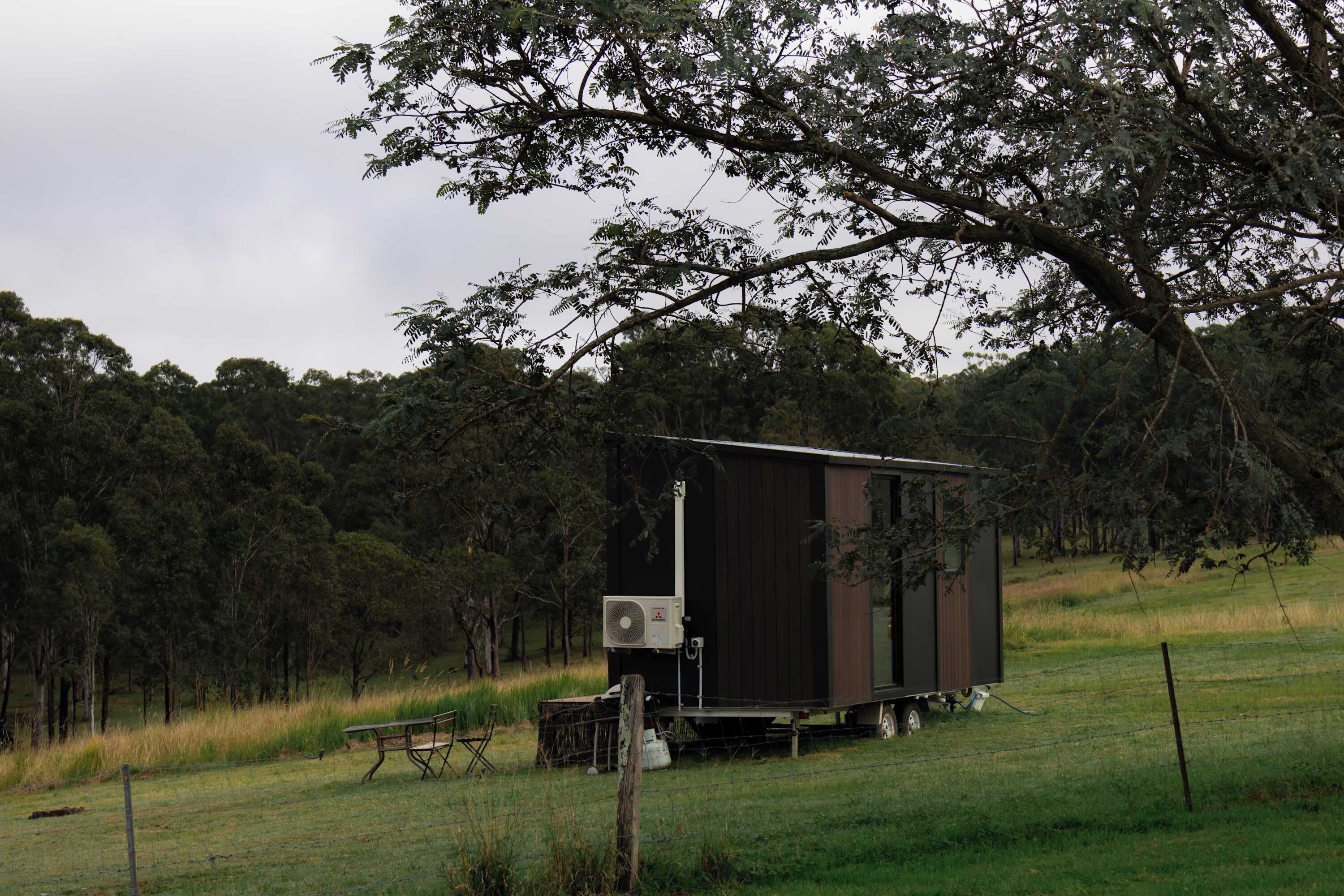 A small, dark-colored mobile home with a flat roof, situated on a grassy field with trees in the background.