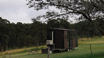 A small, dark-colored mobile home with a flat roof, situated on a grassy field with trees in the background.