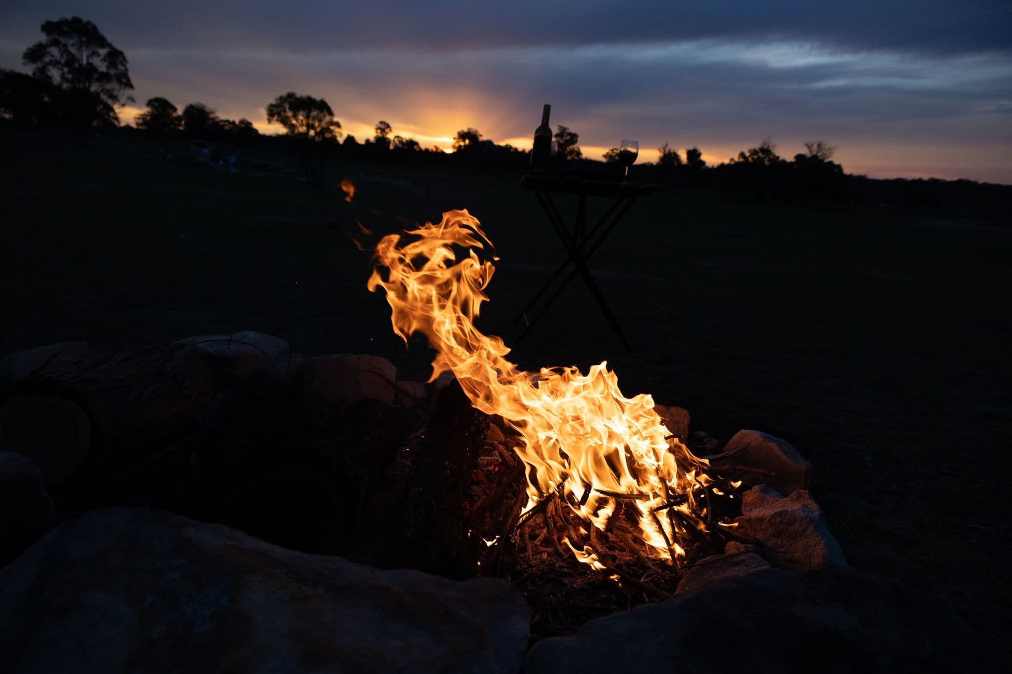 A campfire with logs and a bottle of wine, surrounded by rocks.