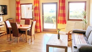 Ein Wohnzimmer mit einer Couch, einem Esstisch mit Stühlen, einem Fernseher und einem Fenster mit Vorhängen.