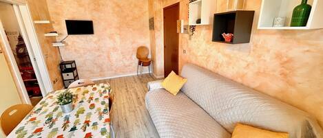 A cozy room with a dining table, a sofa, a TV, and a wall-mounted shelf with decorative items.