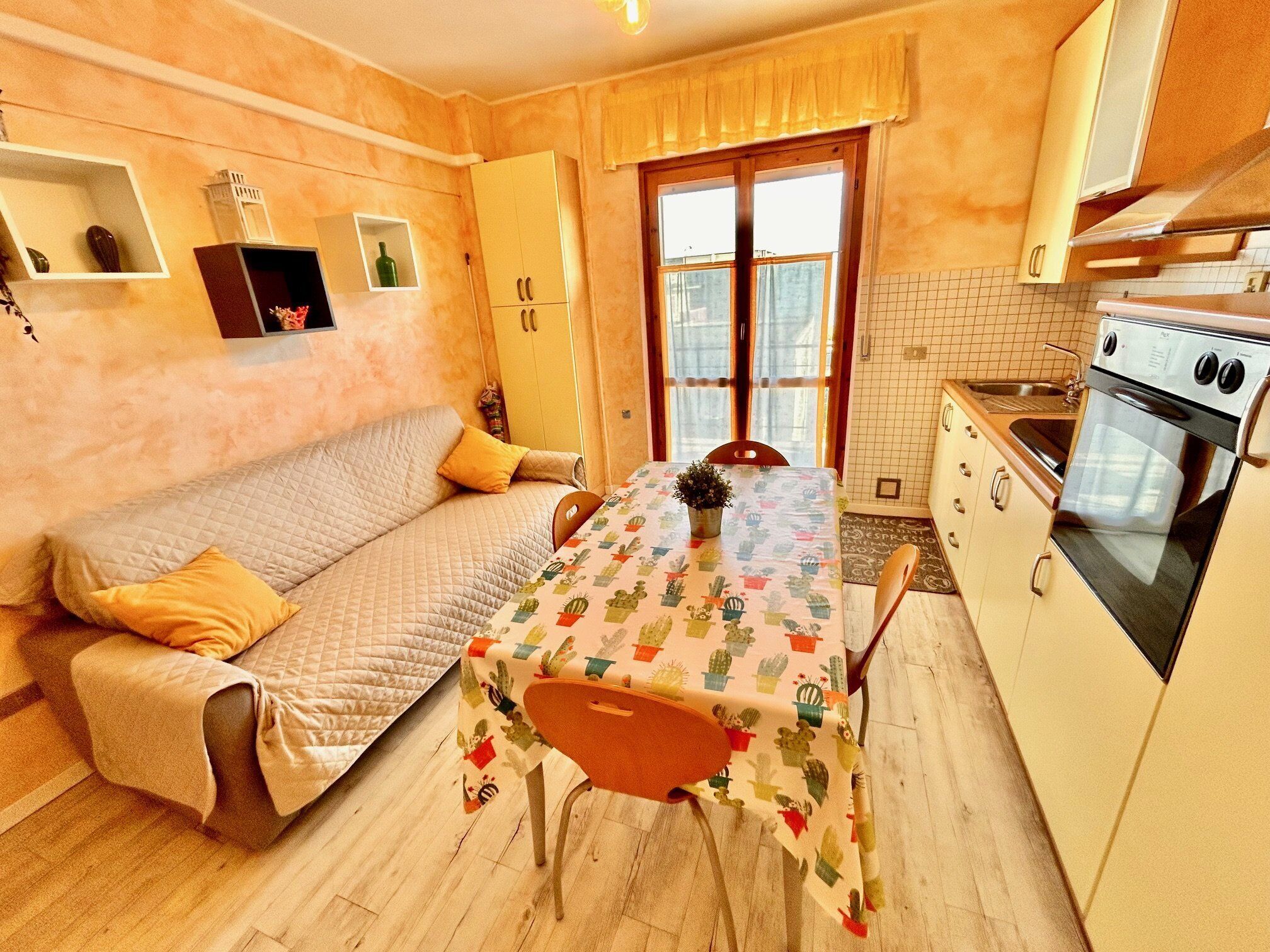A compact living space with a sofa, a dining table with a cactus-patterned tablecloth, a small kitchen with a stove, and a window with a view.
