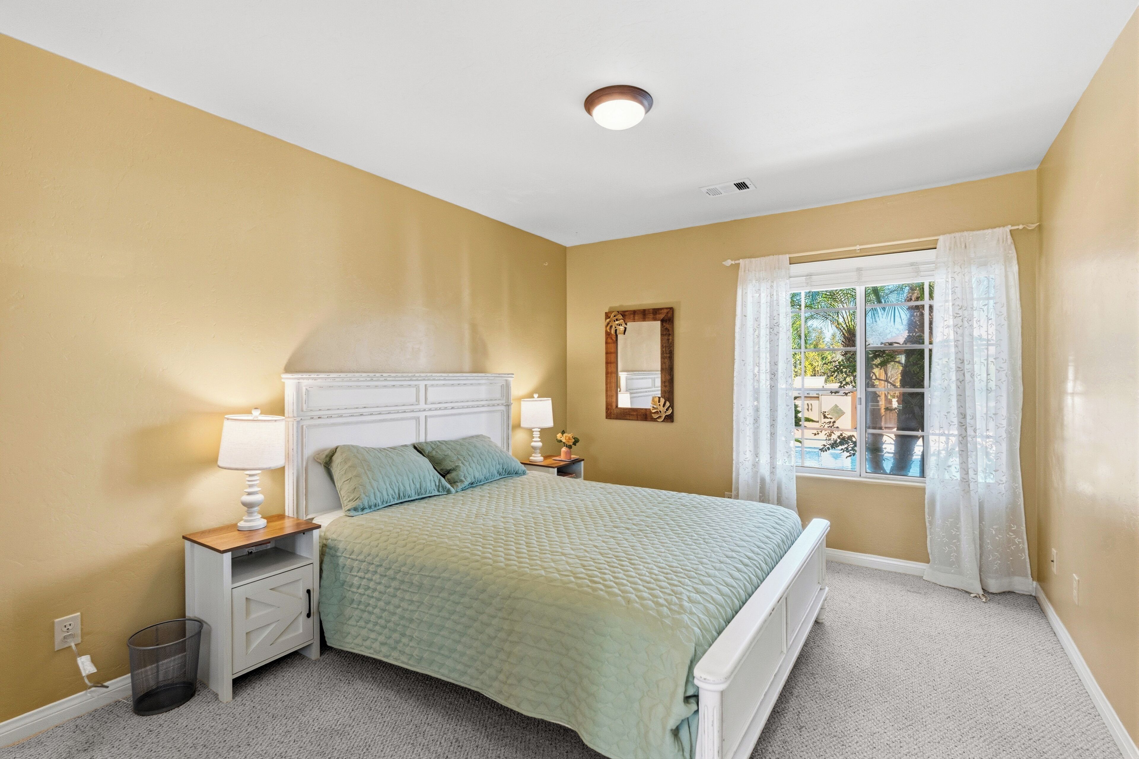 A bedroom with a bed, nightstands, a window with curtains, a mirror, and a ceiling light.