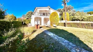 Un bâtiment blanc doté d’un balcon, entouré de haies bien entretenues et surplombé d’un ciel bleu clair.