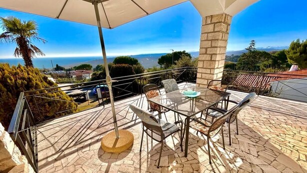Une terrasse extérieure avec un sol en pierre, une table en verre, des chaises en métal et un grand parasol.