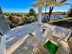 Une terrasse extérieure avec une table et des chaises, un grand parasol et une vue sur la mer et les maisons.