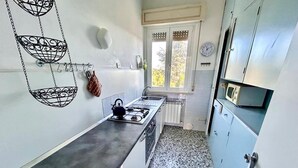 Een smalle keuken met een gasfornuis, hangende keukengerei en een raam met uitzicht op bomen.