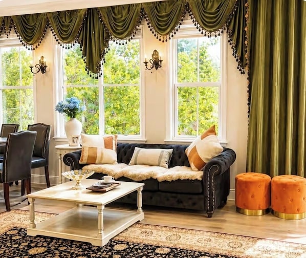 A room with a dark-colored sofa, a white coffee table, and two orange ottomans. There are two windows with green curtains and a view of trees outside.