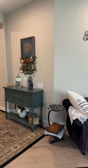 A corner room with a dark green console table, a patterned rug, a dark sofa with a white pillow, and a small side table with a lace cover.