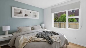 Un dormitorio con cama, lámparas de noche, una ventana con vistas a follaje y decoración en la pared.