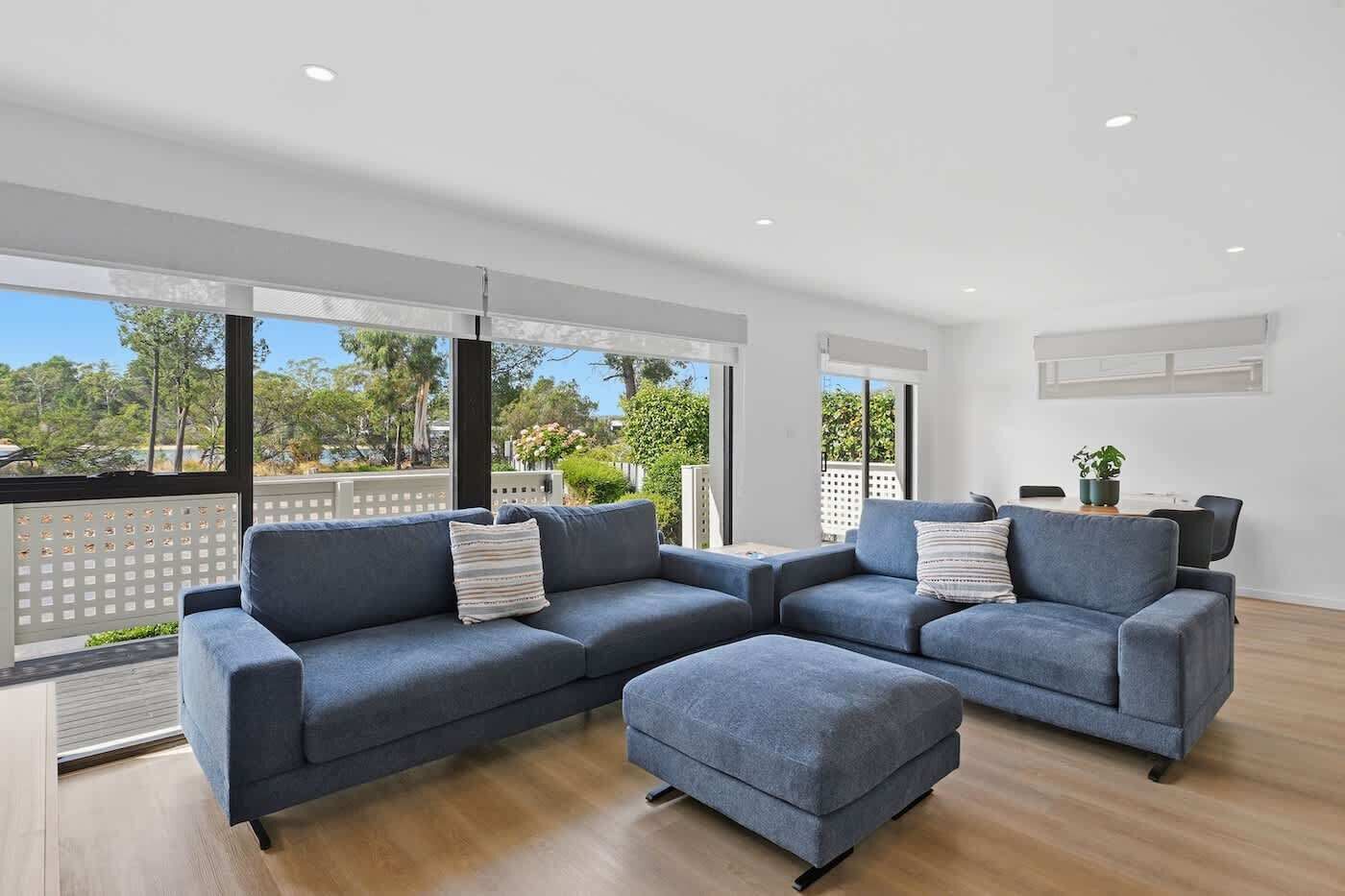 A modern living room with a sectional sofa, a small ottoman, and a dining table with chairs. Large windows offer a view of the outdoors.