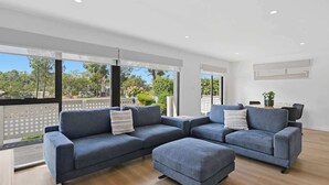 Una sala de estar moderna con un sofá seccional, un pequeño reposapiés y una mesa de comedor con sillas. Amplias ventanas ofrecen vistas al exterior.
