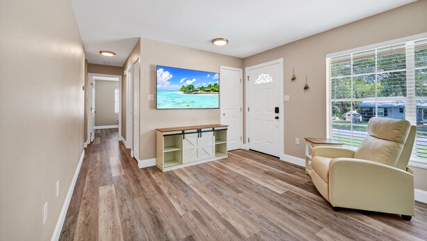 Una habitación con piso de madera, un sillón color beige, un televisor en la pared y una ventana con persianas.