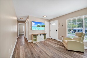 A room with a wooden floor, a beige armchair, a TV on the wall, and a window with blinds.