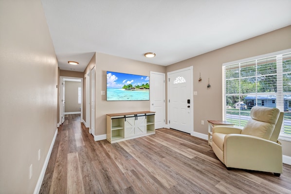 A room with a wooden floor, a beige armchair, a TV on the wall, and a window with blinds.