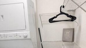 A laundry area with a front-loading washer and dryer, a broom, a trash bin, and wire shelves with hangers. - Cozy 2B2B in Downtown PHX • Near Airport • Perfect Work or Getaway• (Phoenix)