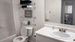 A bathroom with a toilet, a sink, a mirror, and shelves with towels and toiletries. - Cozy 2B2B in Downtown PHX • Near Airport • Perfect Work or Getaway• (Phoenix)