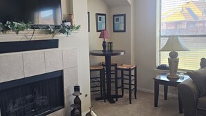 A living room with a fireplace, a television, a dining area with chairs, and a large window with blinds. - Fresh Baked Cookies /Brownies with  check in from Sweet Vegan Cookie Company (Mesquite)