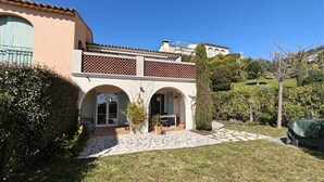 Ein Haus mit einer Terrasse, einem überdachten Sitzbereich im Freien und einem Garten mit verschiedenen Pflanzen.