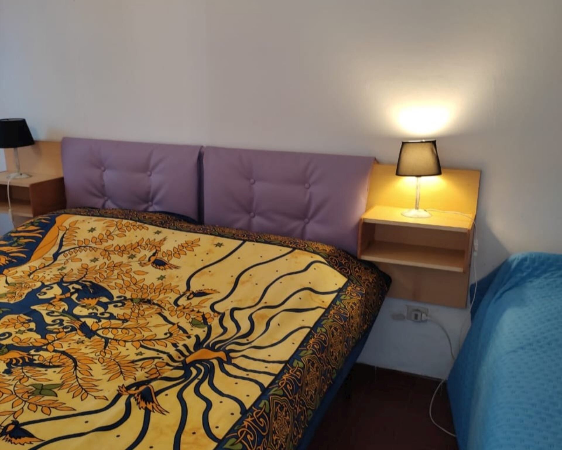 A bedroom with a bed, a nightstand with a lamp, a purple headboard, and a wall socket.