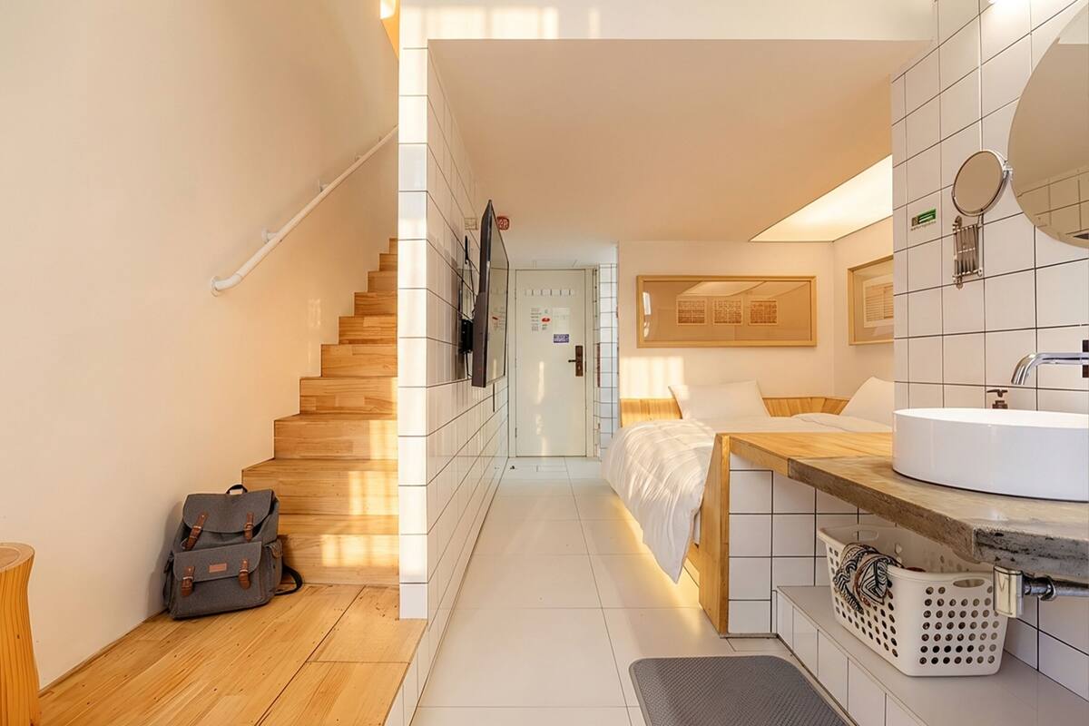Une chambre d’hôtel avec un escalier en bois, un lit avec une literie blanche, une salle de bain avec un lavabo et un miroir, et un petit coin bureau.