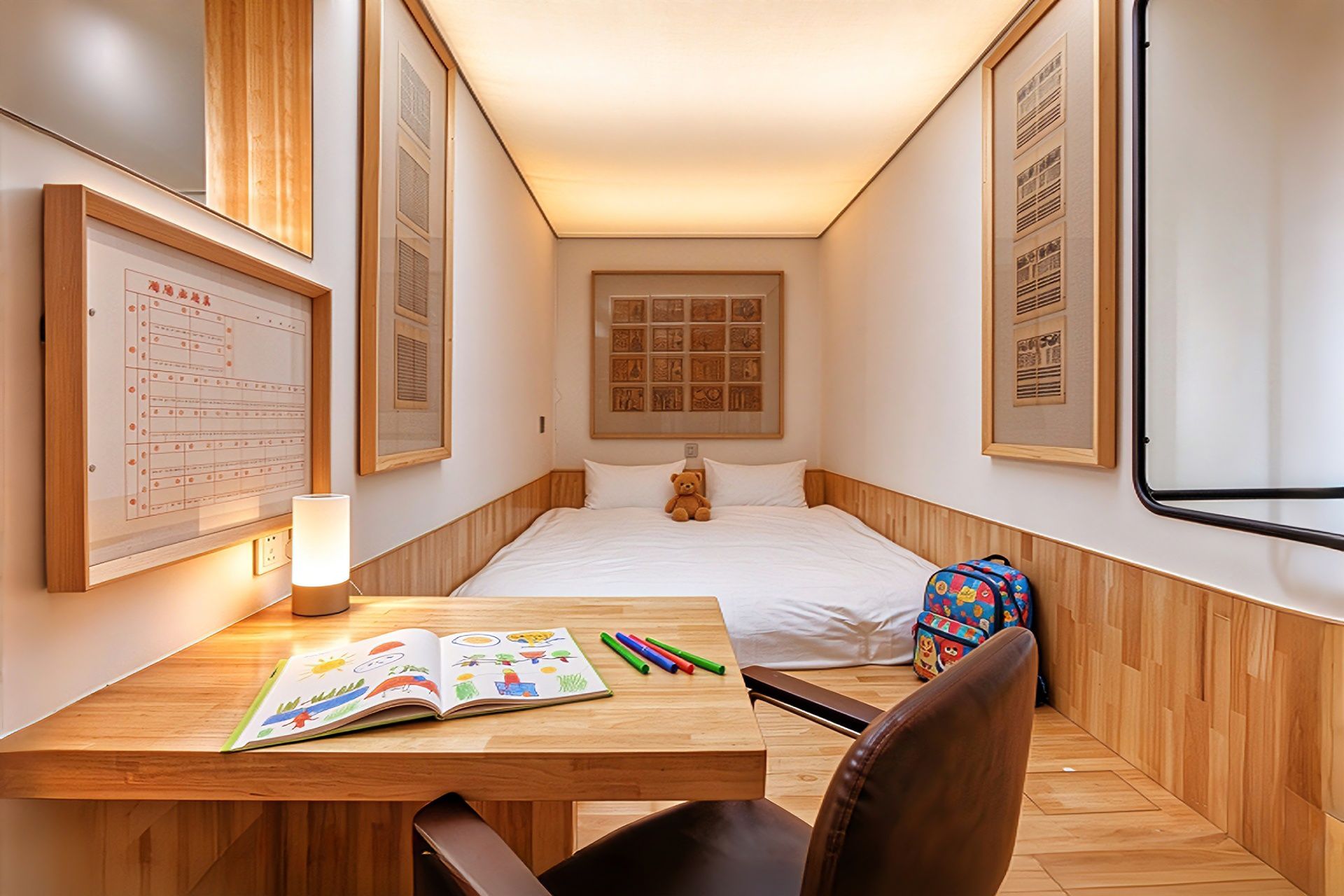 Une chambre d’hôtel avec un lit, un bureau avec un livre et des stylos, un ours en peluche et une fenêtre.