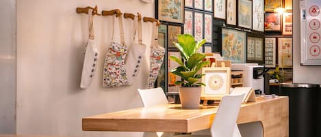 A cafe interior with a wooden table, white chairs, a potted plant, and a wall adorned with framed artwork and hanging bags.