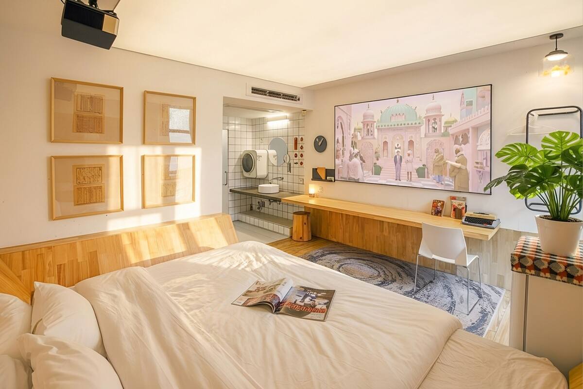 Une chambre d’hôtel avec un lit, un bureau, une chaise, une plante en pot et un grand tableau encadré au mur.