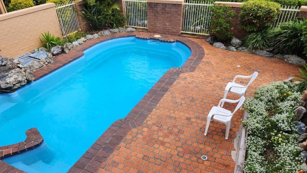Una piscina con borde de ladrillo, rodeada por un patio de ladrillo y una silla de plástico blanco.