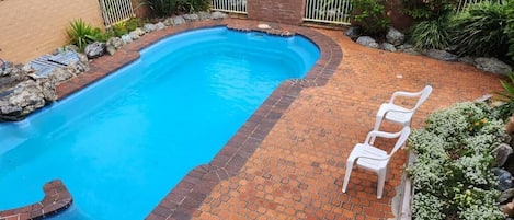 Una piscina con borde de ladrillo, rodeada por un patio de ladrillo y una silla de plástico blanco.