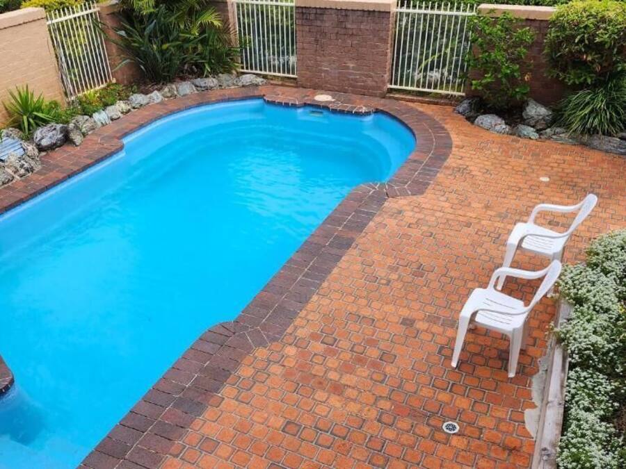 A swimming pool with a brick border, surrounded by a brick patio, and a white plastic chair.