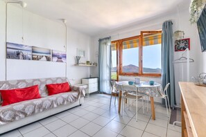A living room with a sofa, a dining table, chairs, a window with a view of mountains, and a ceiling light. - Le Lecce 34, Capoliveri (Capoliveri)