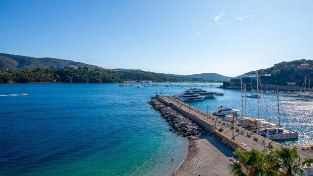 Un puerto costero con barcos amarrados a un muelle de piedra, rodeado de colinas y agua azul cristalina.