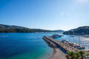 Un port de plaisance situé sur la côte, avec des bateaux amarrés le long d’une jetée en pierre, entouré de collines et bordé d’une eau d’un bleu limpide.