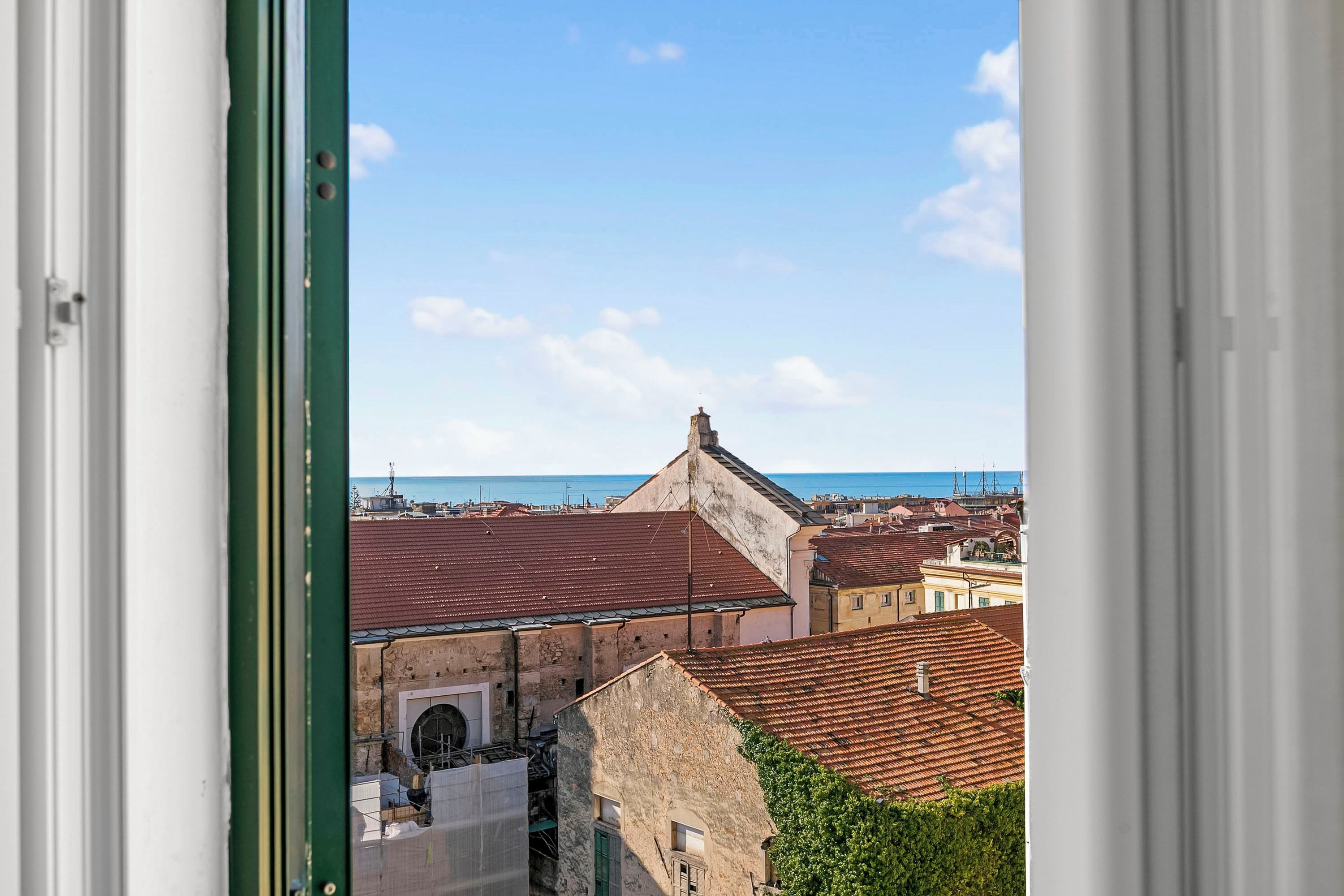 A view through a window of a building with a stone facade, a red-tiled roof, and a glimpse of the sea in the background.