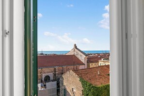 A view through a window of a building with a stone facade, a red-tiled roof, and a glimpse of the sea in the background. - EA Blue Marin Apartment With A/C (Sanremo)