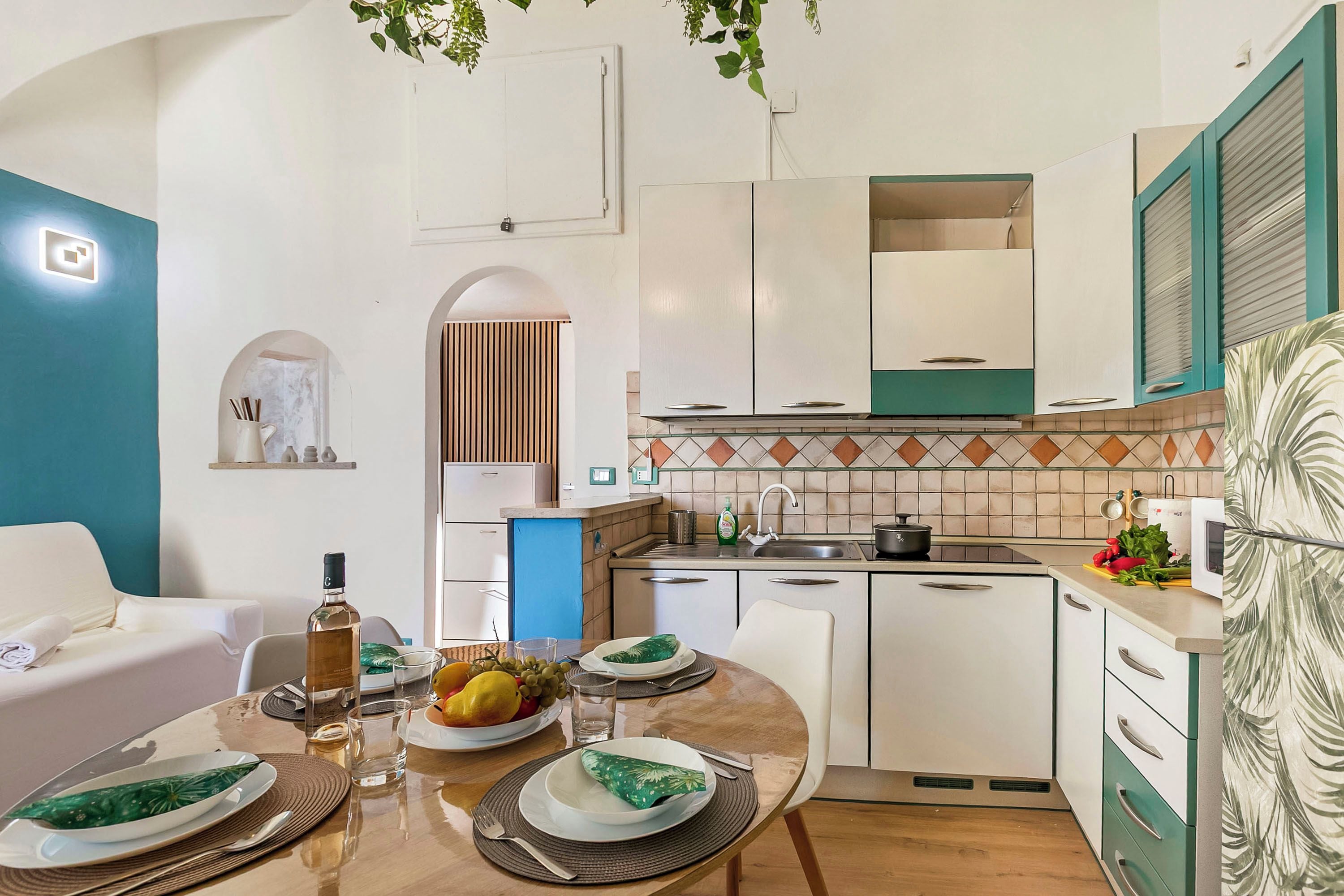 A modern kitchen with white cabinets, a dining table set with plates and cutlery, and a countertop with a kettle and fruit.
