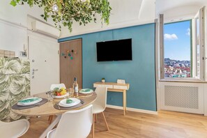 A modern dining area with a round table, white chairs, a flat-screen TV, and a view of a cityscape through a large window. - EA Blue Marin Apartment With A/C (Sanremo)