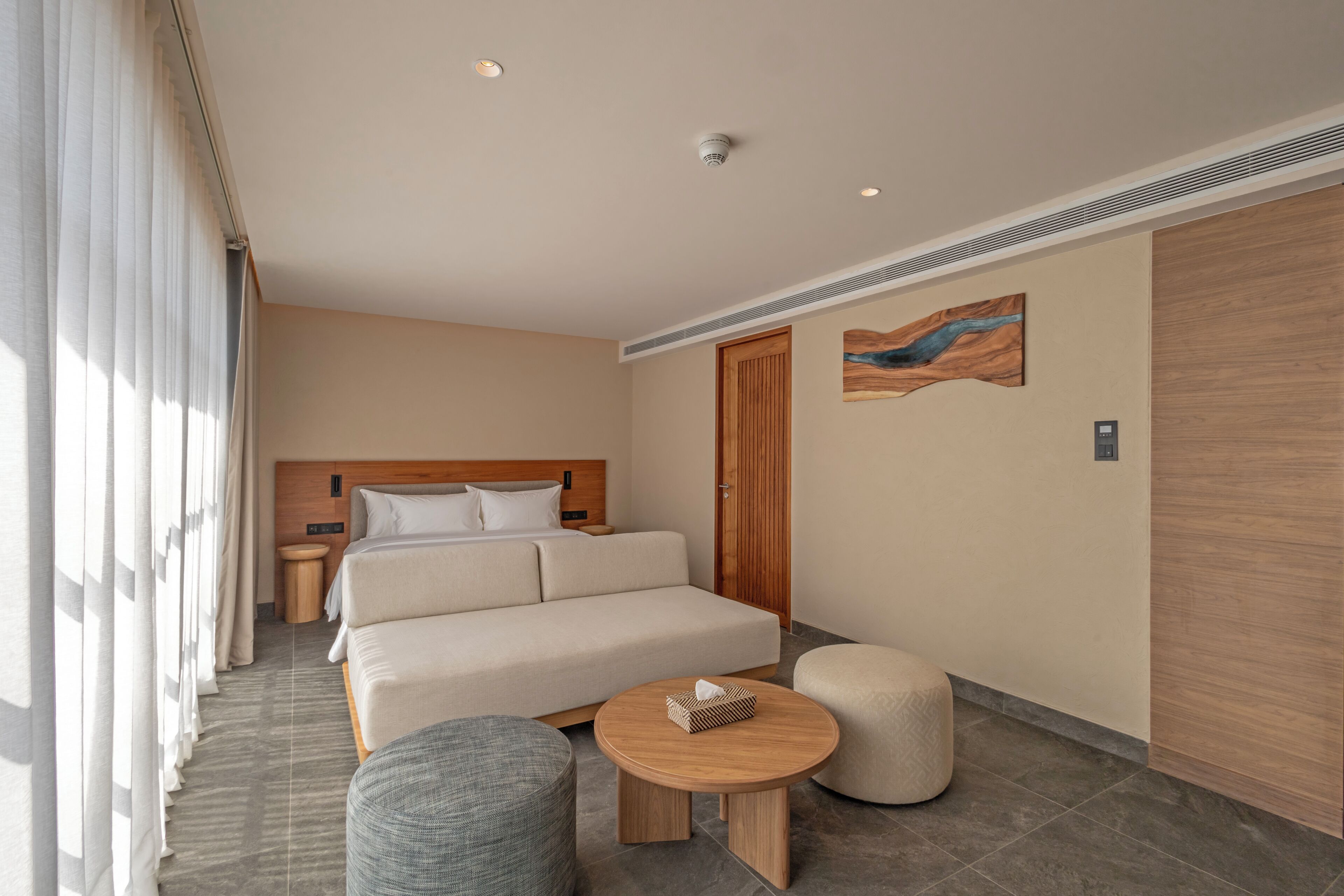 A hotel room with a bed, a round wooden table, a round ottoman, a nightstand, and a wall-mounted artwork.