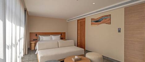 A hotel room with a bed, a round wooden table, a round ottoman, a nightstand, and a wall-mounted artwork.