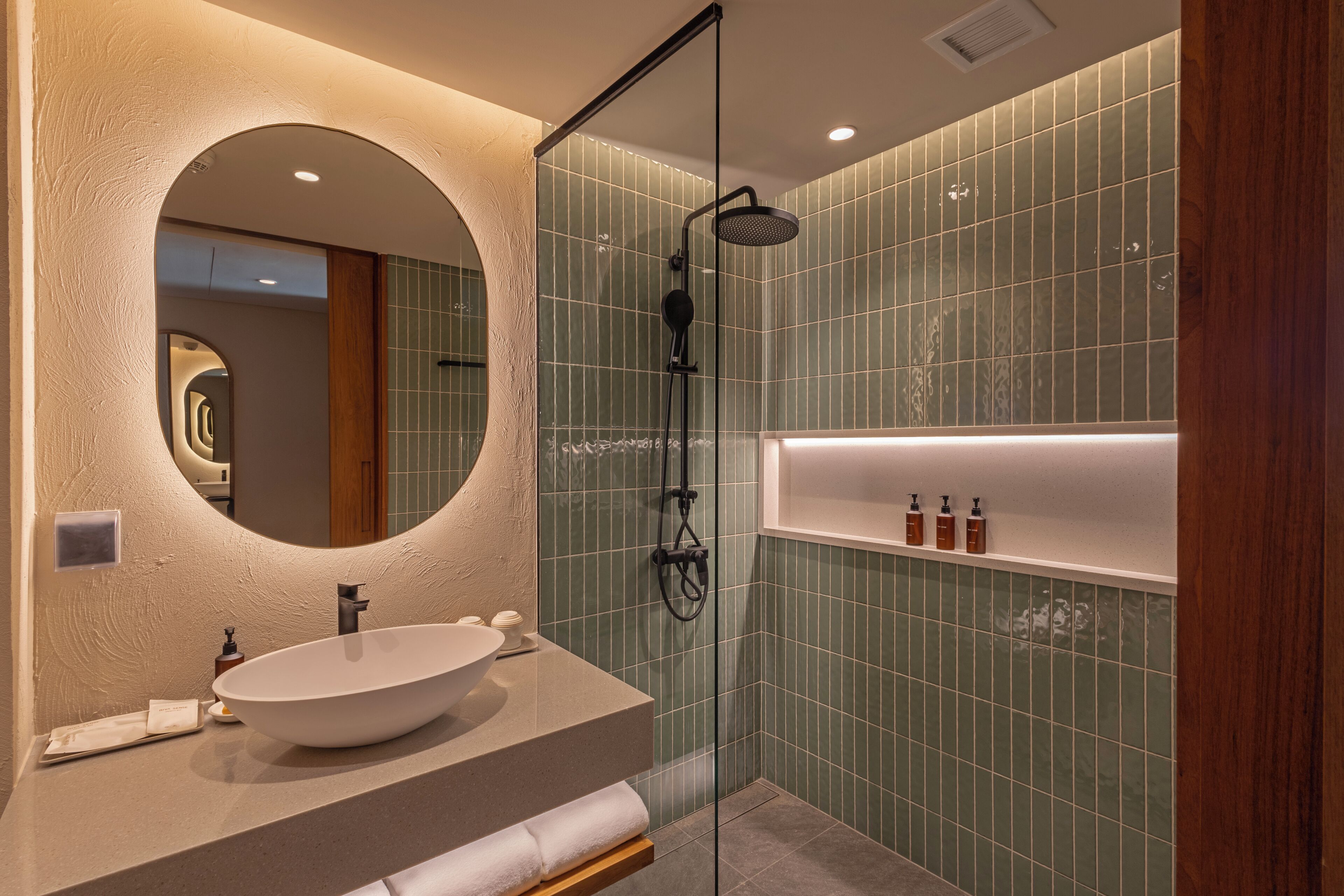 A modern bathroom with a large oval sink, a round mirror, a glass-enclosed shower, and a shelf with toiletries.