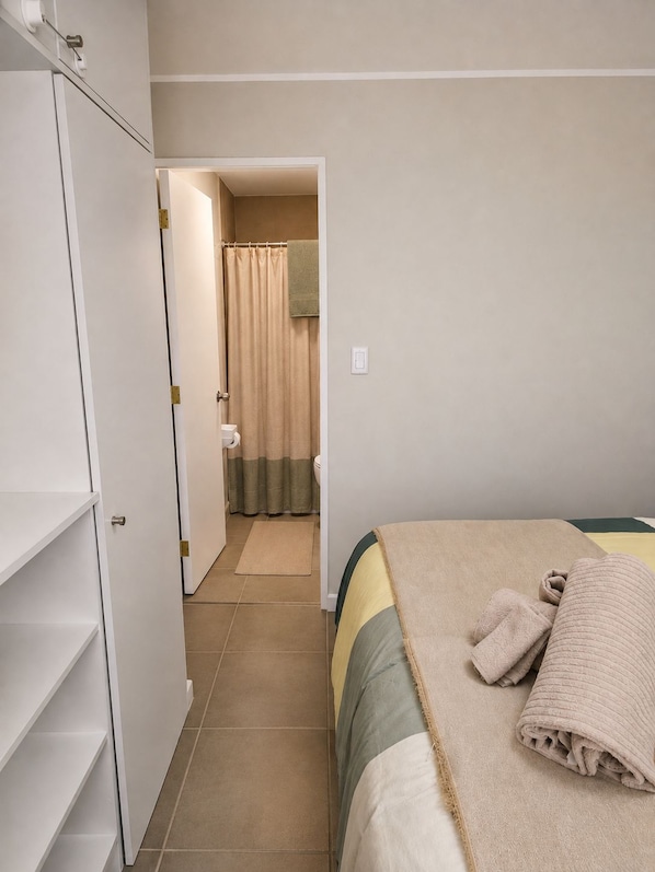Un dormitorio con cama, armario, baño con cortina de ducha y una toalla sobre la cama.