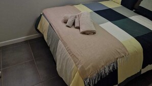 Une chambre à coucher avec un lit, une fenêtre avec des stores, un luminaire au plafond et un sol carrelé.