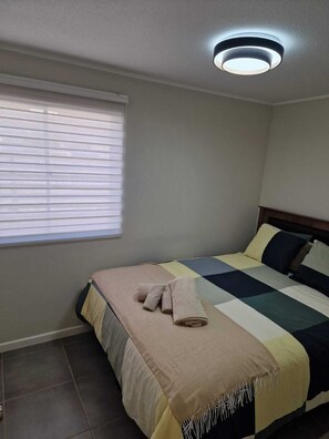 Une chambre à coucher avec un lit, une fenêtre avec des stores, un luminaire au plafond et un sol carrelé.